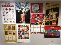 Wide view of nine Coca-Cola posters pinned on a wall showing various vintage and promotional themes, including bottle caps, cans, calendar tiles, and illustration of a woman and child.