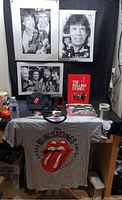Photo showing the gray T-shirt with red tongue logo, posters, cap, book, clock, and other items arranged on a table and wall.