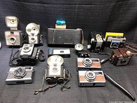 Full lot of 10 vintage cameras arranged on black fabric background with various flash attachments and cases.