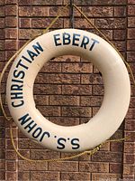 Front side of the ring showing the blue text 'S.S. John Christian Ebert' on white hard plastic, hanging against brick wall