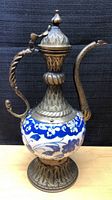 Front view of the entire pitcher showing the ceramic body with blue and white floral design and detailed brass neck, handle, spout and lid showing Ottoman style motifs.