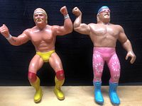 Two WWF wrestling action figures side by side on wooden surface, Hulk Hogan in yellow and red, Dino Ventura in pink and blue.