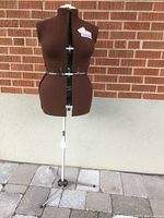Front view of brown fabric covered adjustable dress form mounted on chrome tripod stand outdoors.