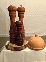 Front view of three wooden salt and pepper grinders on a natural wooden platform next to terracotta garlic roaster with lid.