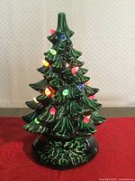 Full view of the green ceramic Christmas tree with lit multi-colored lights on a red cloth surface against a pale wallpaper.