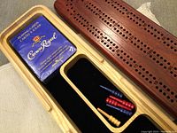 Cribbage board showing hole scoring for pegs, pack of Crown Royal playing cards, and wooden carry case interior with cushioning and peg compartments.