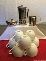 Italian espresso maker, stainless steel creamer, sugar server, and pile of 8 white coffee cups arranged on red cloth and white backdrop.