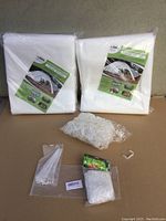 Two folded Flomar Floating Row Covers in original unopened packaging along with loose white plastic clips, a small bag of white zip ties, and a package of trellis netting laid out on a brown surface.