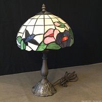 Photo showing the stained glass lamp with floral and hummingbird design on the shade, metal base, and cord visible