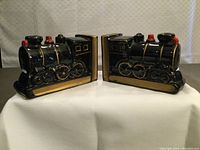 Pair of black ceramic locomotive train engine book ends with red and gold accents shown front-facing, placed on white cloth.