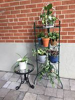 Four-tier green metal plant stand loaded with assorted live and faux potted plants next to a small round glass top metal table with one plant on top, set outdoors on a paved surface against a brick wall.