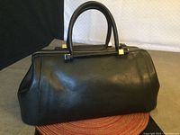 Front view of black leather doctor style handbag with structured silhouette, dual top handles, and gold-tone hardware.