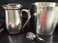 Photo showing the stainless steel Oneida water jug with handle and lid, the tall stainless steel ice bucket with handles, and the clear glass bottle stopper with grape design inside on a metal base.