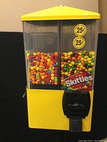 Front close-up of the machine showing two compartments filled with colorful candy and Skittles branding on one dispenser. Yellow top and body with two 25 cent price stickers.