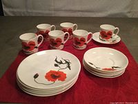 Photo showing 5 lunch plates, 6 coffee cups, and 5 saucers (one saucer is holding a cup) laid out on a red cloth. The plates and cups have a red and black poppy flower design on white china.