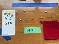 Full view of the pearl necklace laid out flat alongside a red pouch and a card showing the lot number, with a green label marked '14K' indicating gold content.