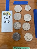 7 Eisenhower dollars and 1 Kennedy half dollar laid out on wooden surface with auction lot card
