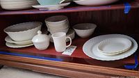 Multiple Arcopal white milk glass dinnerware pieces including stacked plates, bowls, a covered sugar bowl, and mug inside a wooden cabinet shelf.