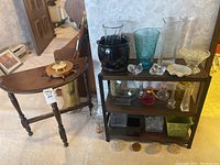 Overall view of wooden half-moon side table with barometer on top, small wooden shelf next to table holding various glass vases and flower frogs.