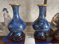Front view of the matching pair of blue and gold cloisonné vases on wooden bases with a floral wallpaper background.