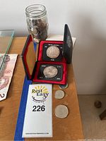 Photo showing two silver Canadian dollar coins in display cases along with loose smaller coins and a jar filled with pennies on a wooden table.