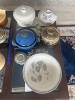 Set of clay pots with lids in white, blue, brown colors and a large signed bowl with leaf design and plate underneath on a wooden table