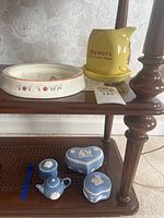 Wide view of the shelf showing Dewar's jug, Toy Town dish, and Wedgewood blue jasperware ceramic boxes and miniature teapot.