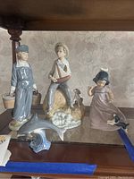 Four LLADRO ceramic figurines displayed on glass shelf: boy with buckets, boy with sailboat and dog, girl with dog, dolphin on waves.