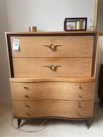 Front view of tallboy dresser showing wood veneer, two top drawers with unique circular ring brass handles, and four curved drawers below with small brass knobs. Legs have brass tips.