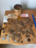 Wide shot showing a collection of old coins scattered on a wooden table, a decorative tin container, and two old paper bills including a visible 100 franc note.