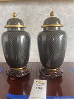 Pair of black cloisonné urns with gold fish scale pattern and lids, sitting on rosewood stands, shown from the front.