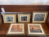 Five framed art prints placed on a wooden surface, showing variety of scenes and frame styles.
