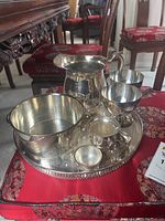 Silver plated pitcher, small pot, two stemmed cups, smaller cup and round tray displayed on a red fabric surface.