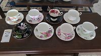 Eight teacups and saucers arranged on table, showing variety of floral patterns and styles including a distinctive black floral set.