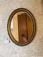 Front-facing photo of the oval mirror showing the gold ornate frame against floral wallpaper.