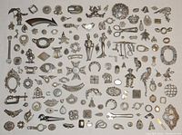 Top-down photo showing a large assortment of over 140 unfinished pewter jewelry pieces laid out in rows on a white surface including figurines, geometric shapes, pins, arrows, and brooches with visible old glue residue.