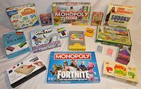 Photo showing group of sealed and used teen/adult games including boxed Monopoly City, Fortnite Monopoly, various card games, puzzle games like Sudoku DVD, Apples to Apples, and others.