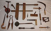 Overview of large assortment of antique hand tools laid out on white fabric including clamp, hand drill, sickle, spokeshave, calipers, hand stapler, and others.