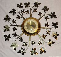 Full view of the sunburst-style brass wall clock with metal leaf decorations radiating from the central clock face lying on a white sheet.