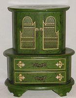 Front view of green armoire jewelry box showing the two cabinet doors with lattice and floral gold detailing and lower two drawers with floral motifs and metal handles.
