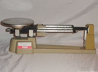 Side view of the entire triple beam balance scale showing beige metal body, circular weighing platform on left, triple sliding beams on right, and brand label on base.