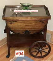 Front view showing walnut tea wagon, carved side panel and large spoked wheel on right, glass tray on top with decorative bowls.