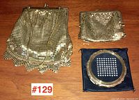 Set of three vintage ladies accessories on wooden surface: one circular compact with mirror and two small gold mesh purses with metal frames and clasps.