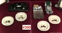 Photo with black, silver, and green vintage model cars, metal cigarette case and ashtray, and four small ceramic plates with vintage vehicle illustrations on a red surface.