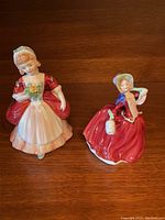 Front view of two Royal Doulton figurines depicting women in red and white dresses with floral and accessory details.