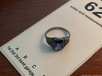 Ring laid on paper background showing full front and side view, prominently featuring large blue emerald-cut stone and marcasite accents on band shoulders.