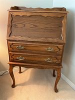 Frontal view of antique style oak drop front desk showing decorative scalloped edges on the drop front and the two drawers with brass handles, all standing on curved cabriole legs.