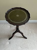 Front view of pedestal table showing scalloped wooden edge, green leather top with gold border and central motif, and three-legged base.