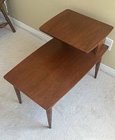 Angled side view showing the two-tier teak side table's shape, wood finish, and tapered legs.