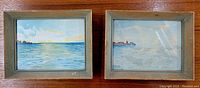 Two framed watercolor paintings showing calm water scenes with sky, one with ship, one with coastline.